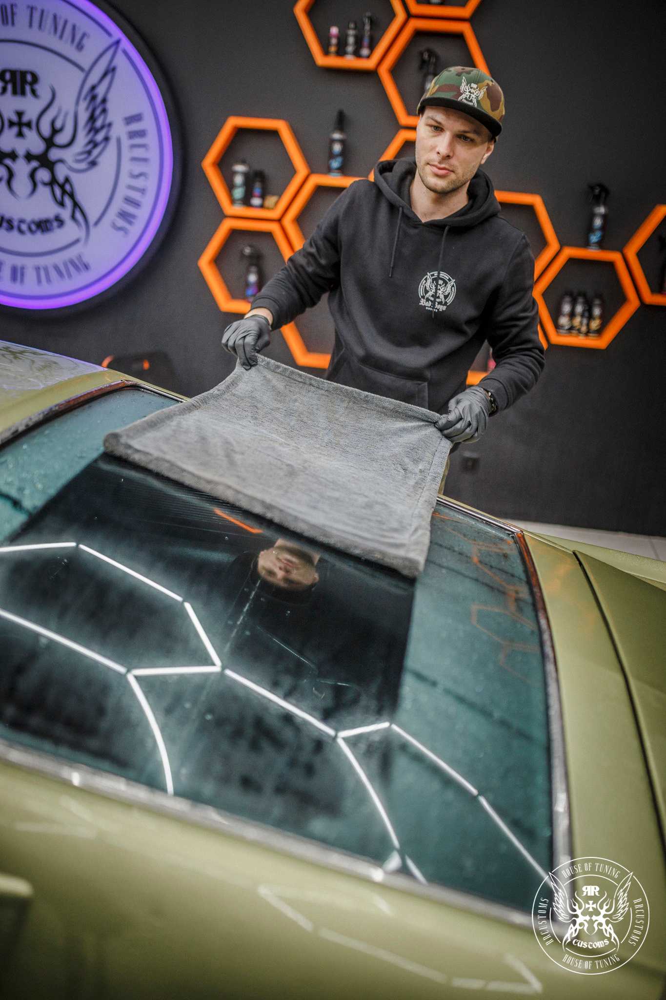Black Master Drying Towel – Detailer tatuado sonriendo mientras seca coche verde militar con la toalla premium negra en estudio hexagonal naranja | Detailing Auto | miio Store