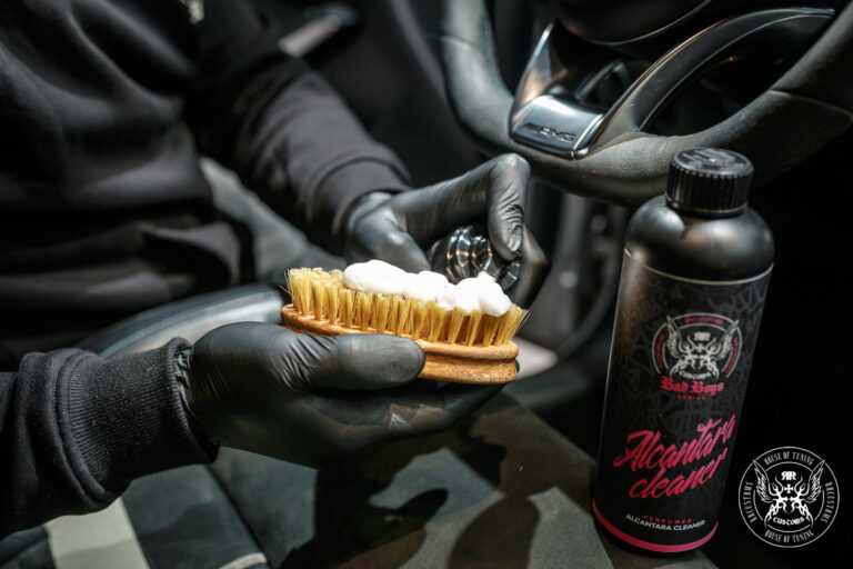 Alcantara Cleaner – Mão com luva preta a usar escova de cerdas naturais para escovar superfície de alcântara, frasco ao lado | Interior Auto | miio Store